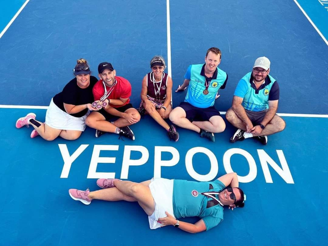 Join the Yeppoon Pickleball Festival in July for nine days of pickleball competition and fun! Here, the medal winners from Hervey Bay sit on the blue court next to the YEPPOON name on court. Everyone is wearing medals and smiling.